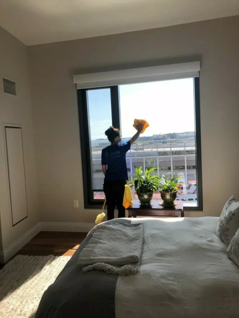 A woman providing maid service in Boston diligently cleans a window in a bedroom.