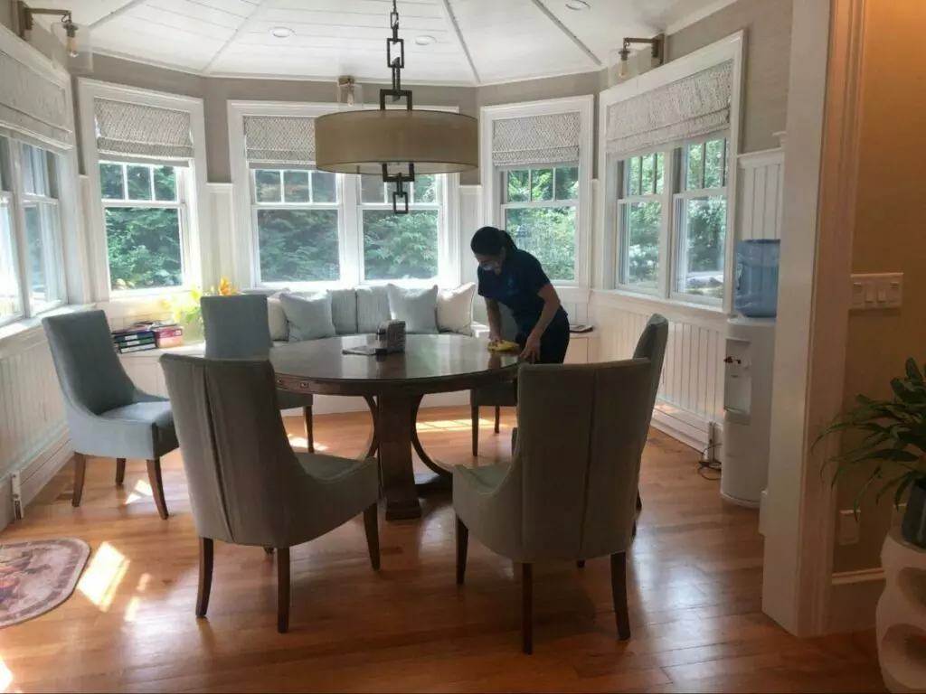 A woman providing cleaning services in a room with a table and chairs.