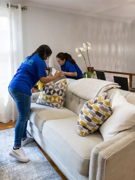 Two women, cleaners from Boston, diligently cleaning a couch in a living room.