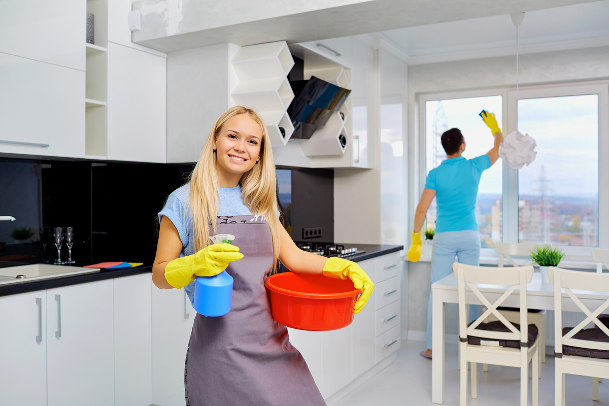 a young couple cleaning the apartment, the wife is carrying a small basin and a sprayer while the husband is wiping the glass window with a sponge