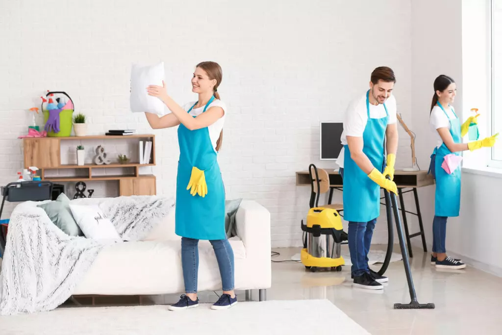 a team of cleaners working together in a living room, with a woman dusting the cushions, another woman wiping the glass window, and a man vacuuming the floor
