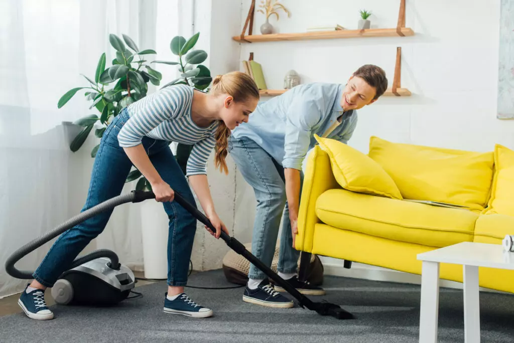 an image of a couple, husband lifts the sofa to make room as wife efficiently vacuums underneath, ensuring a thorough pre-cleaning preparation.
