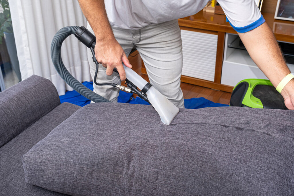 lower body image of a male cleaner using a vacuum cleaner on a gray fabric sofa