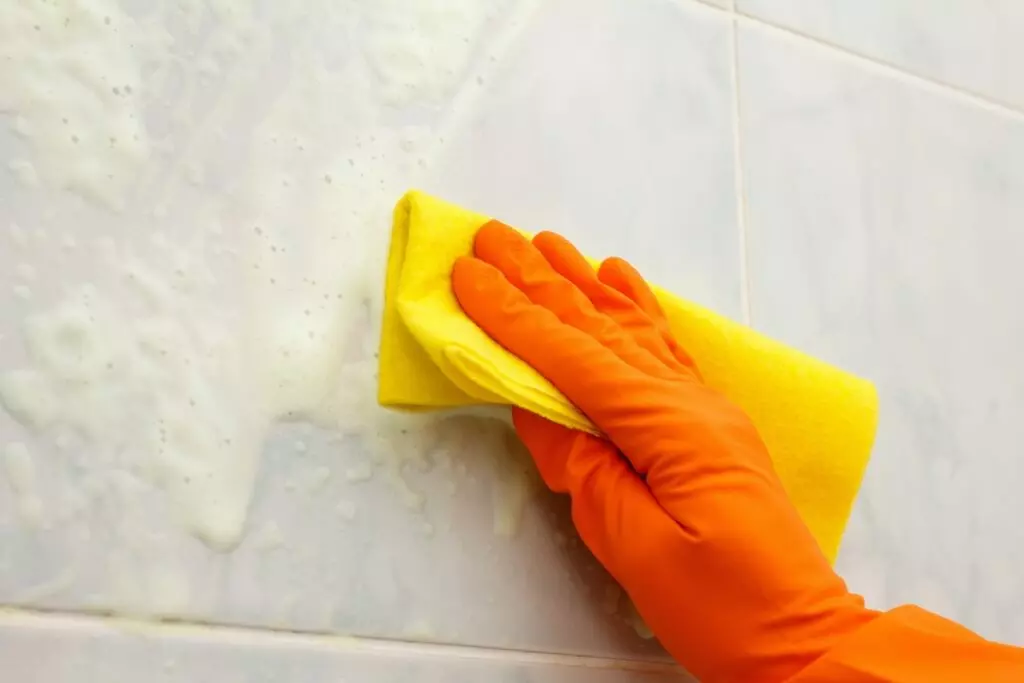 an image of a hand wearing gloves cleans soapy bathroom tiles with a cloth