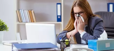 A woman in a business suit is blowing her nose at her desk in the office.