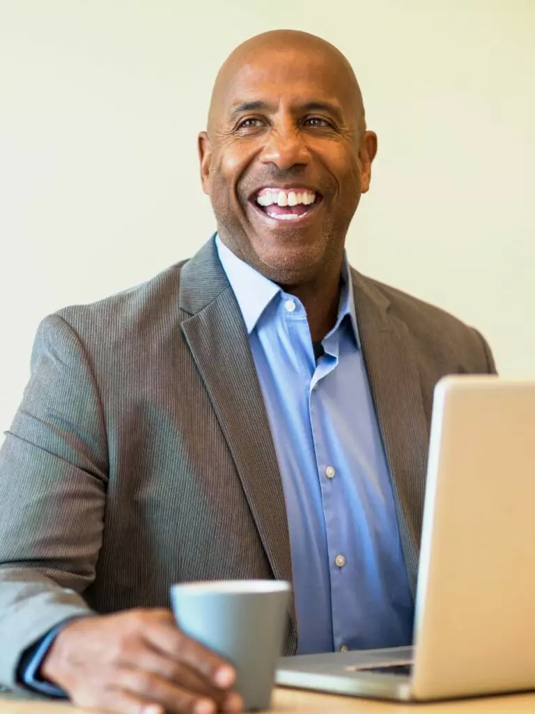 A smiling black man in a suit sitting at a desk working on a laptop.