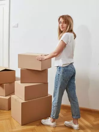 Woman carrying a cardboard box in a room with multiple boxes, possibly involved in house cleaning or organizing.