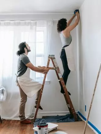 A person standing on a stepladder applying painter's tape to the top corner of a room while another assists by holding the ladder for safety during house cleaning.