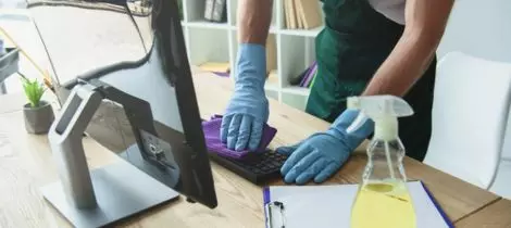 Person cleaning a computer keyboard with a cloth while wearing gloves, using eco-friendly cleaning products.