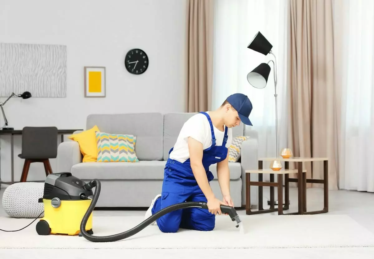 A man in blue overalls providing cleaning services while vacuuming carpet in a living room.
