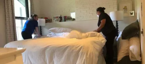 Two people house cleaning by making a bed in a room with brick walls and bedside lamps.