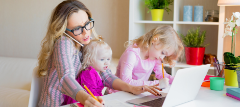 A woman wearing glasses works on a laptop at a desk with two young children who are coloring, in a brightly lit home office designed for spring cleaning planning.