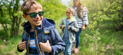 A young boy in sunglasses smiles while hiking in a sunny, wooded area during spring cleaning, with two people walking in the background.