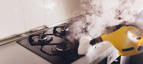 A person wearing a glove uses a yellow steam cleaner from their kitchen cleaning checklist to clean a kitchen stove emitting steam.